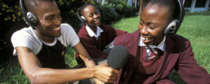 A young man interviews a young girl in her school uniform. There is another schoolgirl in the background. Everyone is wearing headphones. The young man is holding a microphone and a recording device.