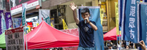 A young man stands on a table in a busy market.. He holds a microphone with one hand and his other hand is raised