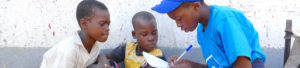 A school pupil dressed in a blue t-shirt and cap bends down to write something on a piece of paper. They are interviewing two younger children who are looking at what is being written with interest.